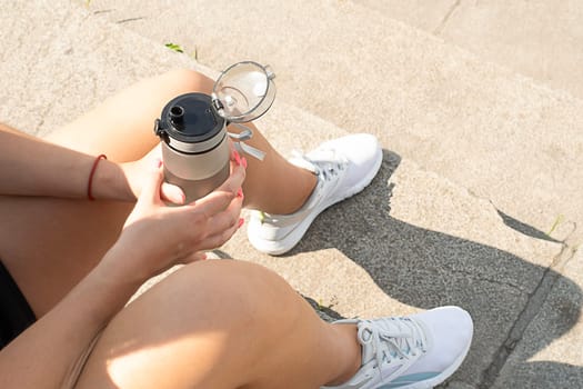 Sports concept. The girl sits on the steps in white sneakers and holds a bottle of water in her hand. Resting and quenches thirst after a workout on the street. No face. Close-up.