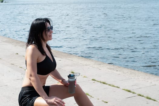 Sports concept. A girl sits on the steps in white sneakers, a T-shirt and shorts and holds a bottle of water in her hand. Resting and quenching thirst after training on the embankment near the river.