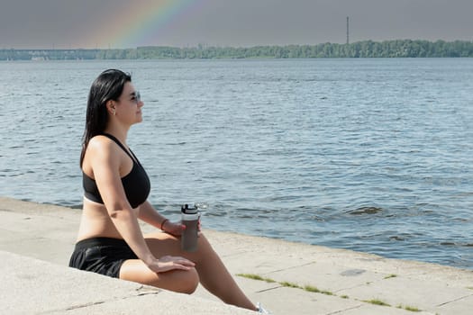 Sports concept. A girl sits on the steps in white sneakers, a T-shirt and shorts and holds a bottle of water in her hand. Resting and quenching thirst after training on the embankment near the river.