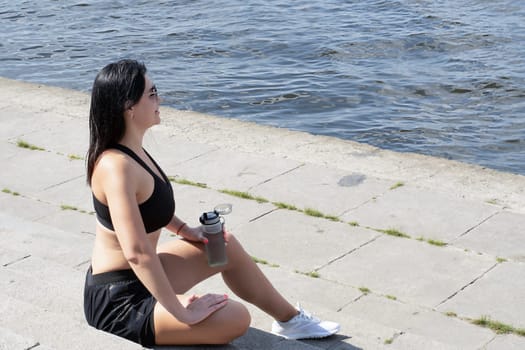 Sports concept. A girl sits on the steps in white sneakers, a T-shirt and shorts and holds a bottle of water in her hand. Resting and quenching thirst after training on the embankment near the river.