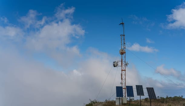 The communication antenna on top of the mountain is powered by solar panels and a small wind turbine, with a cloudy blue sky in the background.