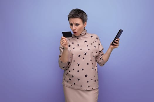 surprised 30 year old european woman with dyed gray short hair in a skirt and blouse looks at the screen of the phone and at the bank card.