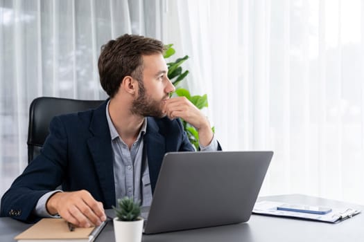 Businessman in black suit working on laptop at his workspace desk. Smart executive researching financial data and planning marketing strategy on corporate laptop at modern workplace. Entity