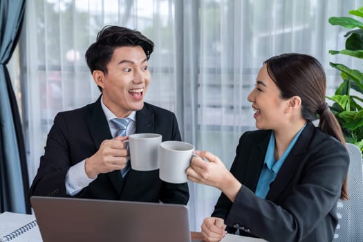 Two Asian office workers taking coffee break together in workplace. Coworkers smiling and socializing while holding cup of coffee adding friendly working environment in corporate workspace. Jubilant