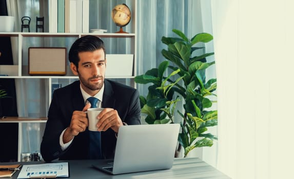 Modern professional businessman at modern office desk using laptop to work with coffee in his hand. Diligent office worker working on computer notebook in his office work space. fervent