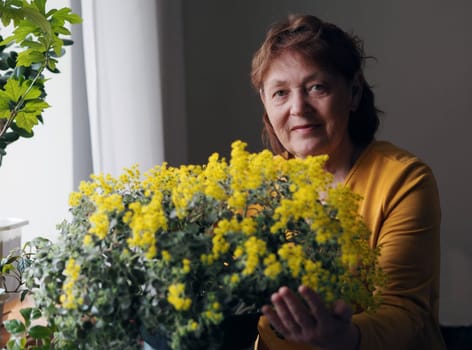 Portrait of an elderly woman in a yellow dress with a blooming houseplant Aichrizon with yellow flowers. Life and favorite hobbies of pensioners.