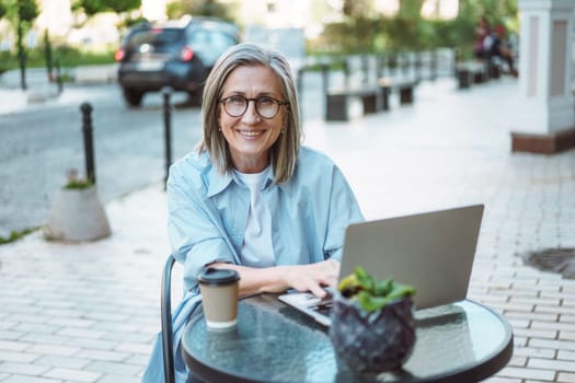 Working from anywhere, smiling and happy mature woman working on laptop from cafe on street. Freedom and flexibility that comes with remote work, allowing individuals to choose their work environment. High quality photo