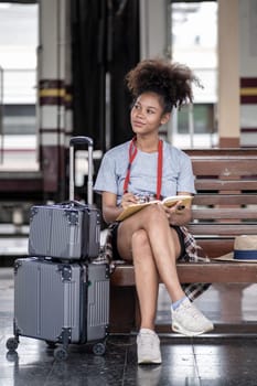 Asian African female tourist traveler holding mobile phone smart phone sitting at train station, Confident smiling teenager girl playing holding book smart phone laptop computer on suit case at station. High quality photo