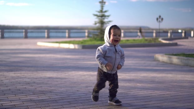 A one-year-old son runs along the paving stones on the embankment. A small child in a sweater with a hood. 4k.