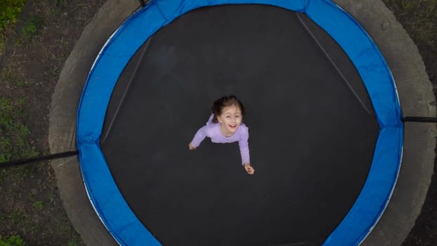 View of the jumping girl from the drone. A little girl is jumping on a trampoline in the yard. The camera moves away. 4k