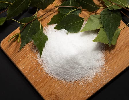 Useful sweetener Xylitol, birch sugar on cutting wooden board,sticks, leaves of birch, dark background. Healthy nutrient, diet. Flat lay. Horizontal plane