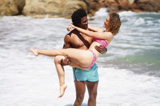 Smiling young multiracial couple in swimwear standing near waving blurred sea water and looking at each other while African American male carrying girlfriend in hands