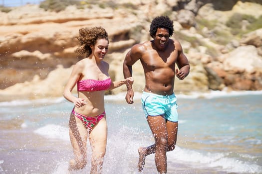 Young multiracial couple in swimwear running in splashing water while looking down and laughing together during sunny day near sea waves