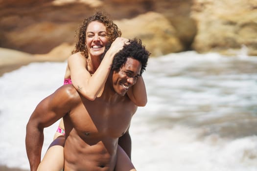 Joyful young African American man giving piggyback ride to cheerful curly haired woman having fun on seashore during sunny vacation laughing and smiling