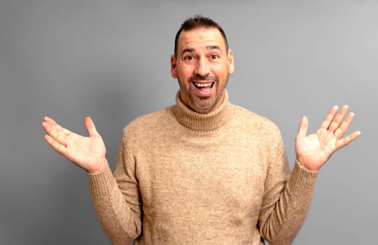 Bearded Hispanic man in his 40s wearing a turtleneck sweater with arms outstretched in an expression of surprise, isolated on gray studio background