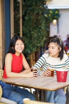 Cheerful young Asian female friends, in casual apparel sitting at table with cold drinks and speaking about plans for future while spending time together in cafe