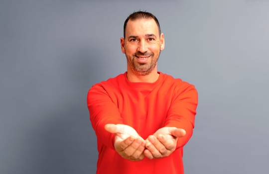 Bearded Hispanic man in his 40s wearing a red jumper offering something imaginary in his clasped hands, isolated on gray studio background