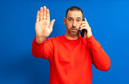 Bearded Hispanic man in his 40s wearing a red sweater making the stop sign while talking on the cell phone visibly serious and angry, isolated on blue studio background