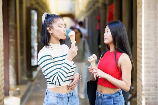 Side view of young Asian girlfriends eating ice cream cone while standing together in street against building