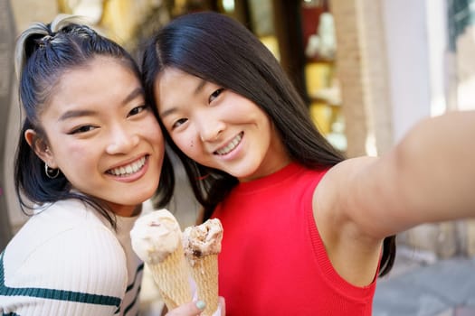 Positive young Asian women smiling happily and taking selfie while eating delicious ice cream cone together in street