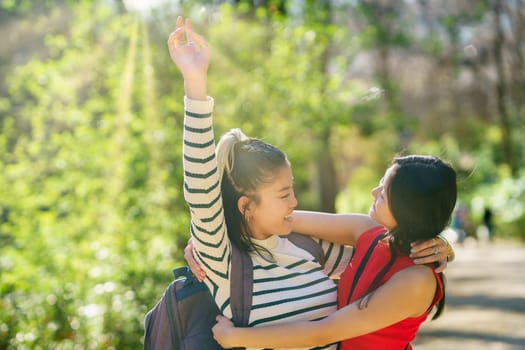 Happy young Asian female travelers in casual outfits, smiling and looking at each other while embracing with raised arm in sunny forest of Alhambra during trip through Granada