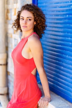 Beautiful young female in red sundress looking away while leaning on stone wall with hand against blue shutters in daylight