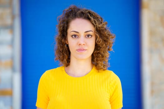 Charming young curly haired female in yellow t shirt looking at camera while standing on city street against blurred background