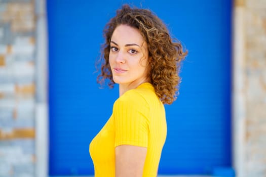 Side view of cheerful young female in yellow t shirt, with curly brown hair looking at camera and smiling against blue wall of shabby building on sunny street