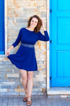 Full body of happy young female in blue dress smiling and looking at camera while touching curly brown hair on paved street