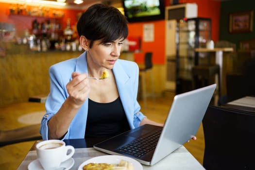 Concentrated young female in blue jacket with short hair, holding snack in spoon and sitting at table with coffee while looking at screen of laptop in restaurant