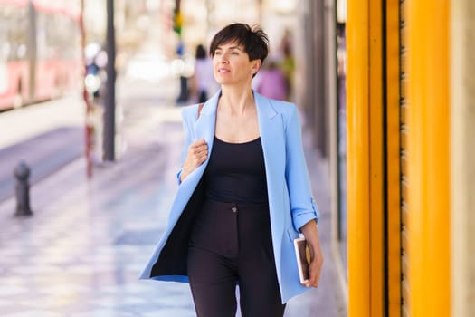 Confident mature woman in formal clothes standing on city street and looking away while holding planner against blurred city background