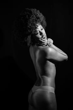 Sensual nude African American female with curly hair looking away over shoulder while standing against black background. Black and white photograph.