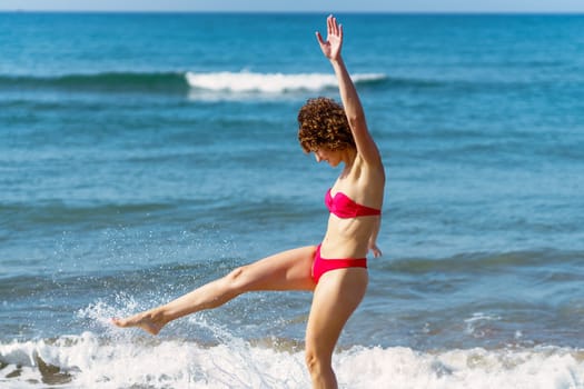 Side view of cheerful young fit female with short curly hair in red swimwear smiling and kicking water while standing on seashore during summer vacation