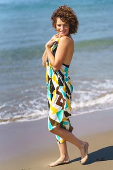 Full body side view of positive young female in colorful dress standing on sandy beach near waving sea in daylight and looking at camera