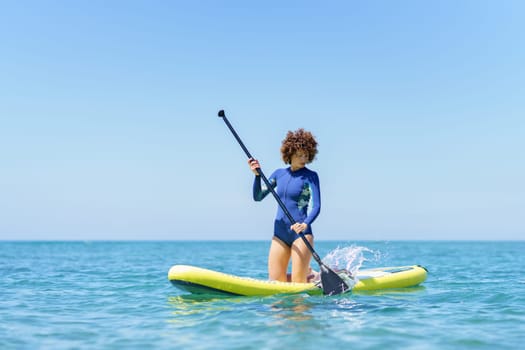 Focused curly hair woman in wetsuit with paddle kneeling on SUP board while paddling on blue sea under blue cloudless sky