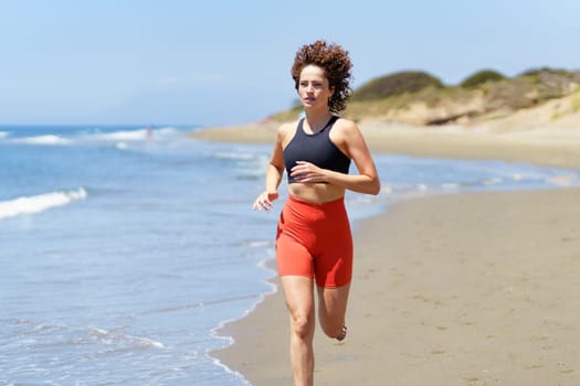 Concentrated barefoot young female in sportswear and with wavy hair looking away, while jogging on sandy beach in daylight near seawater against blurred sky and grassy dunes