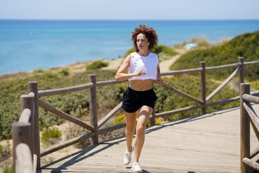 Full body of young sportswoman in activewear with curly hair running on wooden walkway near sea during fitness training