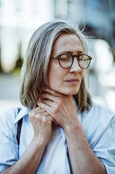 Concept of sickness through mature lady holding throat. Woman appears sick and tired, with weary expression on face. Cold toned color palette, adding to overall sense of illness and discomfort. . High quality photo