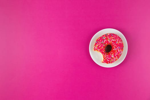 A donut with colored sprinkles lies on a white plate, which stands on a pink background. Concept confectionery, pastries, coffee shop. Flat lay, top view, photo for banner.