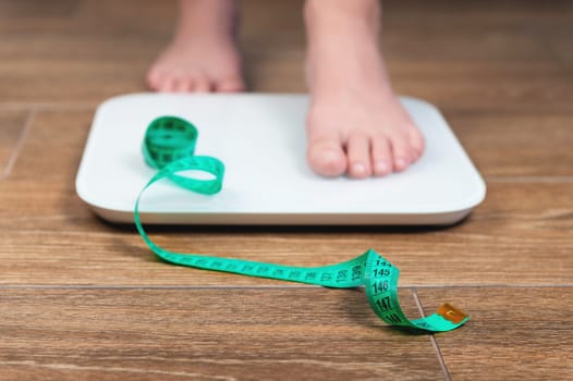 Cropped image of female legs standing on scales on wooden background. Roulette in the foreground. The concept of weight and figure measurement, beauty standards.