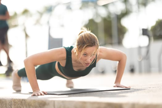 Adult sportive woman with no make up doing push ups on yoga mat outdoors. Mid shot