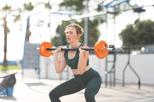 Adult athletic woman squatting with dumbbells in her hands standing on outdoors sports ground. Mid shot
