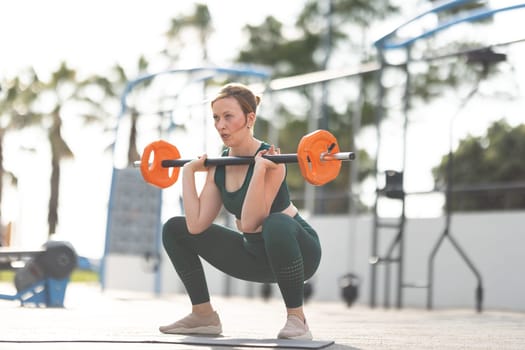 Adult athletic woman squatting with dumbbells in her hands standing on the sports ground. Mid shot