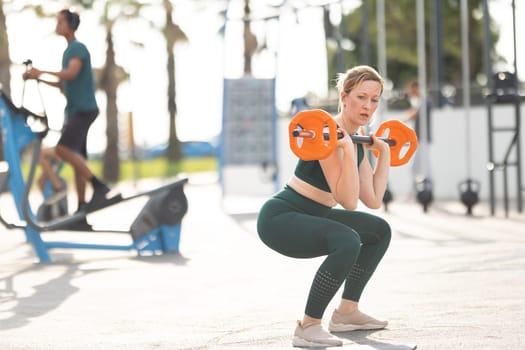 Adult athletic woman with nice body squatting with dumbbells in her hands standing on the sports ground. Mid shot