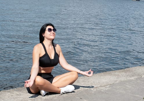 Yoga concept. A beautiful girl in a lotus position sits on the embankment near the river in the street in the fresh air. Meditate with closed eyes.