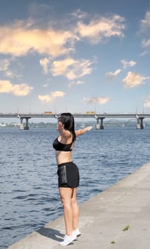 Sport concept. A slender and tall girl in sportswear and white sneakers stands near the river with her arms outstretched to the side and inhales the fresh air.
