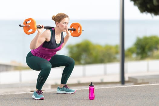 Adult athletic woman squatting with a dumbbell on her shoulders - a plastic bottle of water in front of her. Mid shot