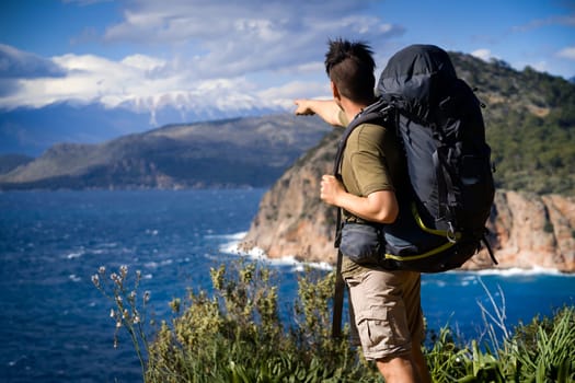 A young man is traveling, trekking and walking along the sea coast on a sunny day with a backpack. The traveler points with his hand to a beautiful mountain on the horizon.