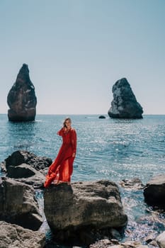 Woman travel sea. Young Happy woman in a long red dress posing on a beach near the sea on background of volcanic rocks, like in Iceland, sharing travel adventure journey