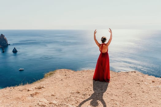 Side view a Young beautiful sensual woman in a red long dress posing on a rock high above the sea during sunrise. Girl on the nature on blue sky background. Fashion photo.